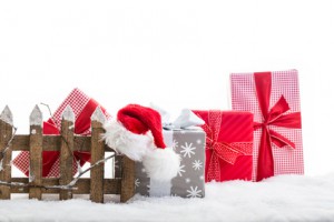 verschiedene Geschenke auf Kunstschnee mit einem Holzzaun auf dem eine Weihnachtsmütze hängt