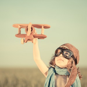 Junge spielt mit einem Holzflugzeug und hat eine Fliegerbrille und Mütze auf