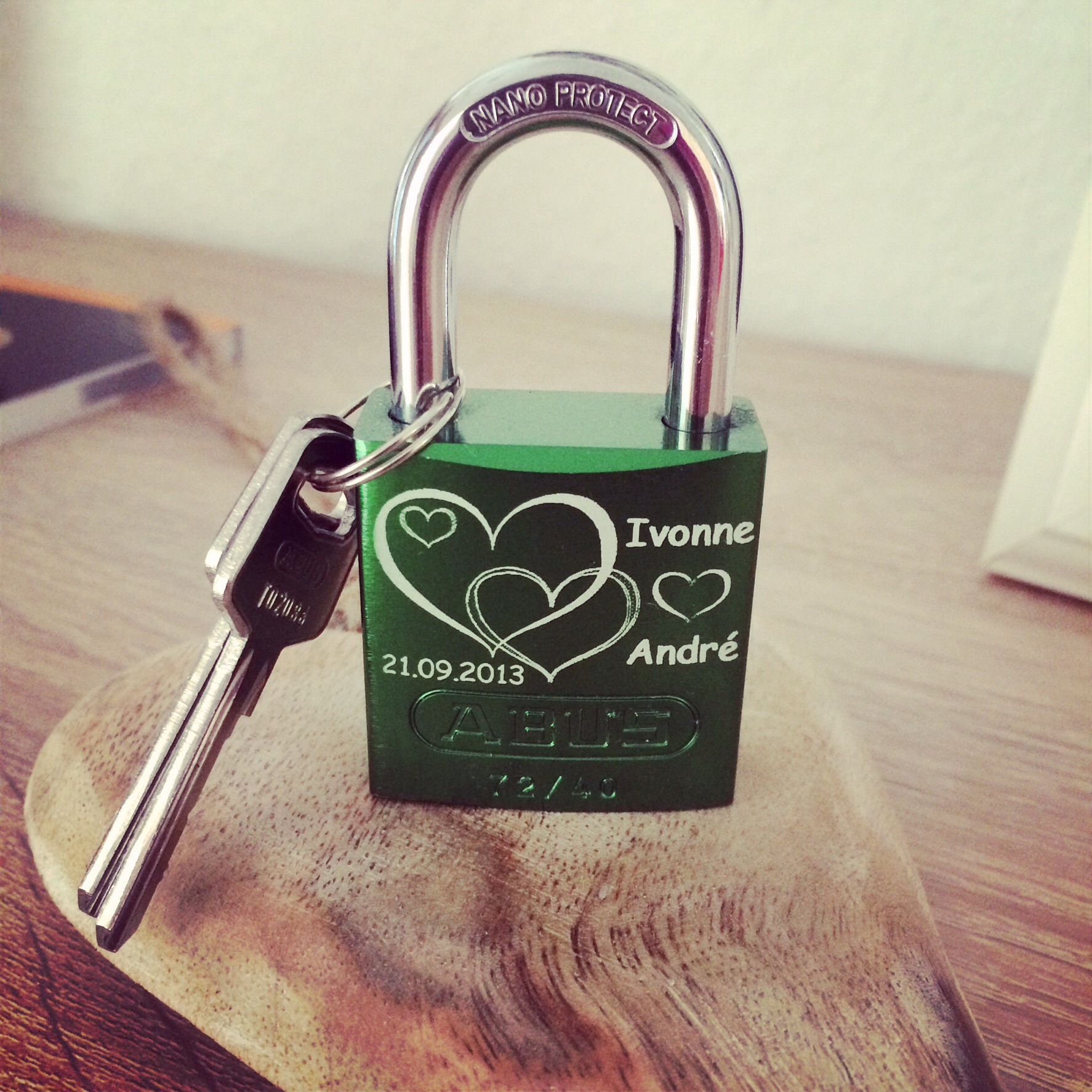 Grünes Liebesschloss mit Gravur und angehängten Schlüsseln auf einem Holzbrett.