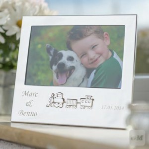 Bilderrahmen in Silber steht auf einer Fensterbank. In dem Bilderrahmen ist ein Bild von einem kleinen Jungen mit einem Hund. Auf der Vorderseite ist unter dem Foto, mittig eine Eisenbahn mit einer Gravur auf der rechten und linken Seite.