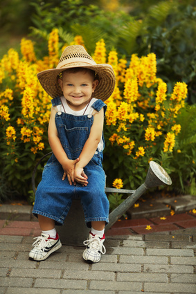 Kind mit Hut sitzt auf Gießkanne vor gelben Blumen
